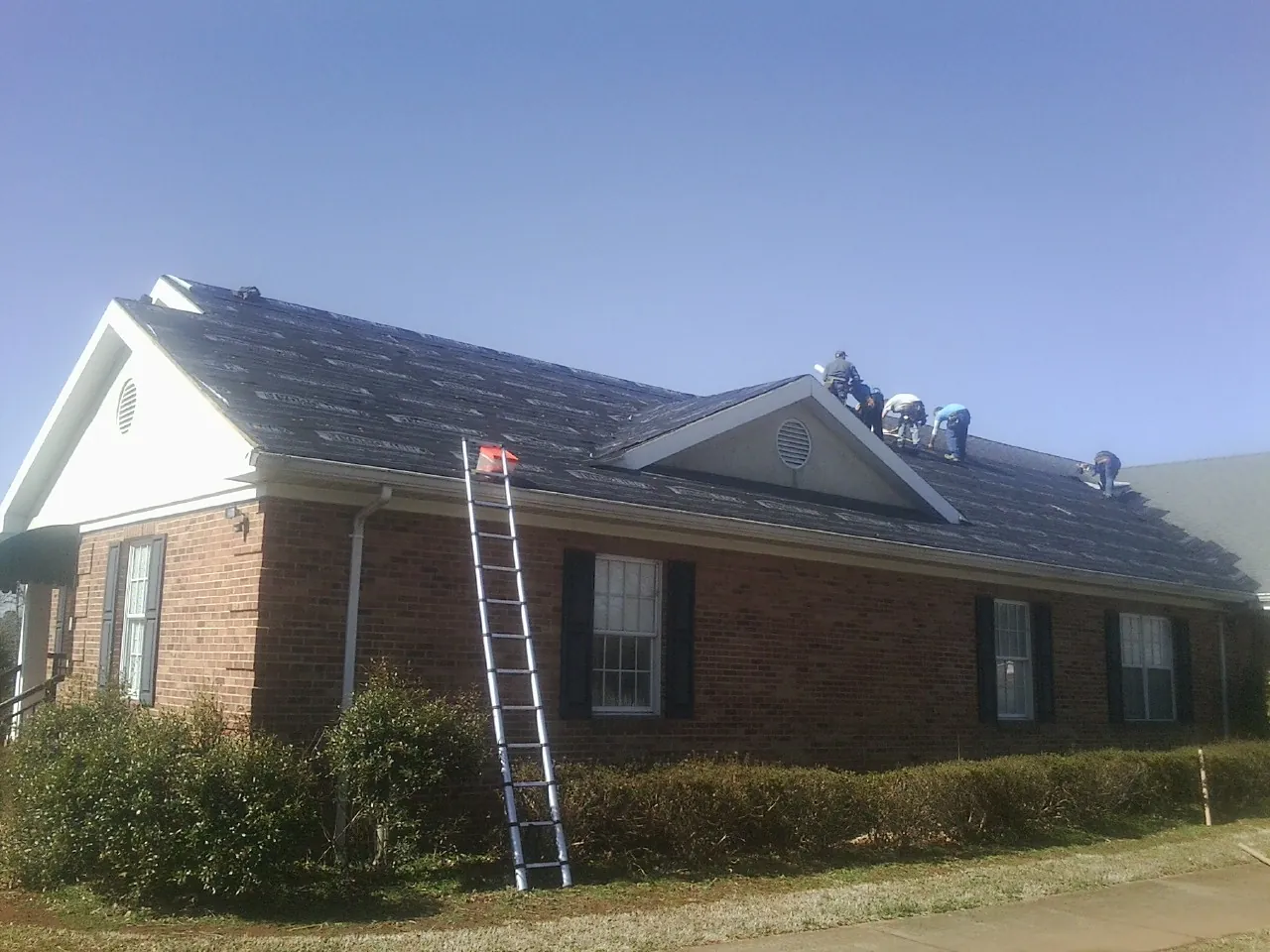 Crew tearing shingles off a brick ranch roof with a ladder set — Emergency Roof Repair work by our crew in Mc Leansville