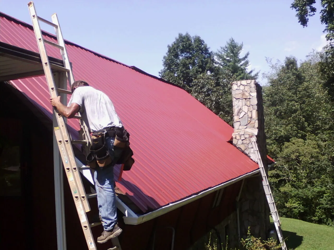 Roofer on a ladder beside a red metal roof and stone chimney — Soffit & Fascia Repair by Roofing Brothers in Mc Leansville